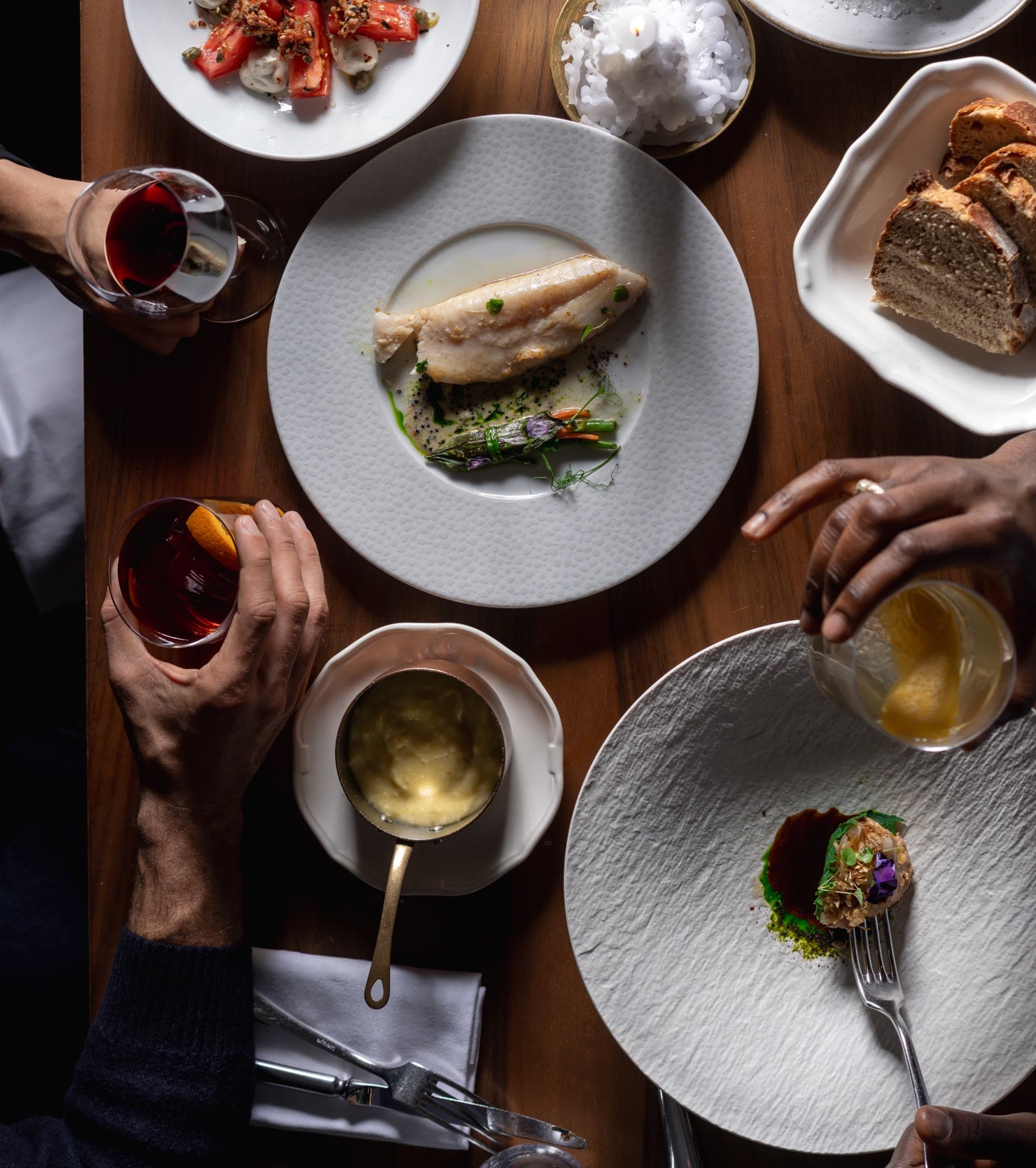 Une table à partager vue du dessus : poisson, tomates, purée, pain et verres de vin entre les mains des convives